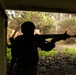 A silhouetted figure in tactical gear and helmet is aiming a rifle from inside a structure toward a lush, tropical outdoor environment. A second figure is partially visible behind them near a wall. The scene appears to depict a military or law enforcement training exercise in a tropical setting.