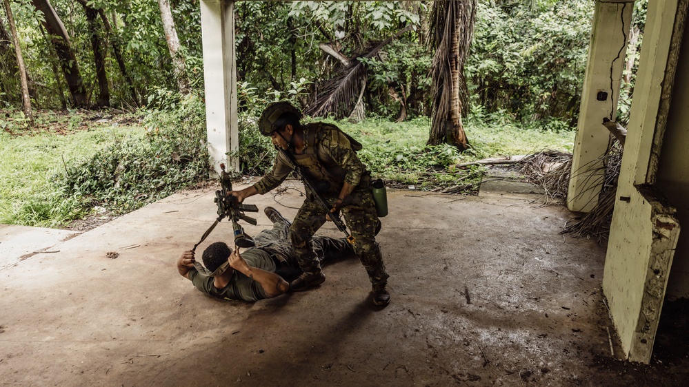 Two individuals in military or tactical gear are engaged in what appears to be a close-quarters combat training drill inside an open-air concrete structure surrounded by dense tropical vegetation. One person in full camouflage uniform and helmet is standing over another person on the ground, with a rifle visible in the scene. The setting suggests a jungle training environment or simulated urban combat exercise.