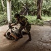 Two individuals in military or tactical gear are engaged in what appears to be a close-quarters combat training drill inside an open-air concrete structure surrounded by dense tropical vegetation. One person in full camouflage uniform and helmet is standing over another person on the ground, with a rifle visible in the scene. The setting suggests a jungle training environment or simulated urban combat exercise.