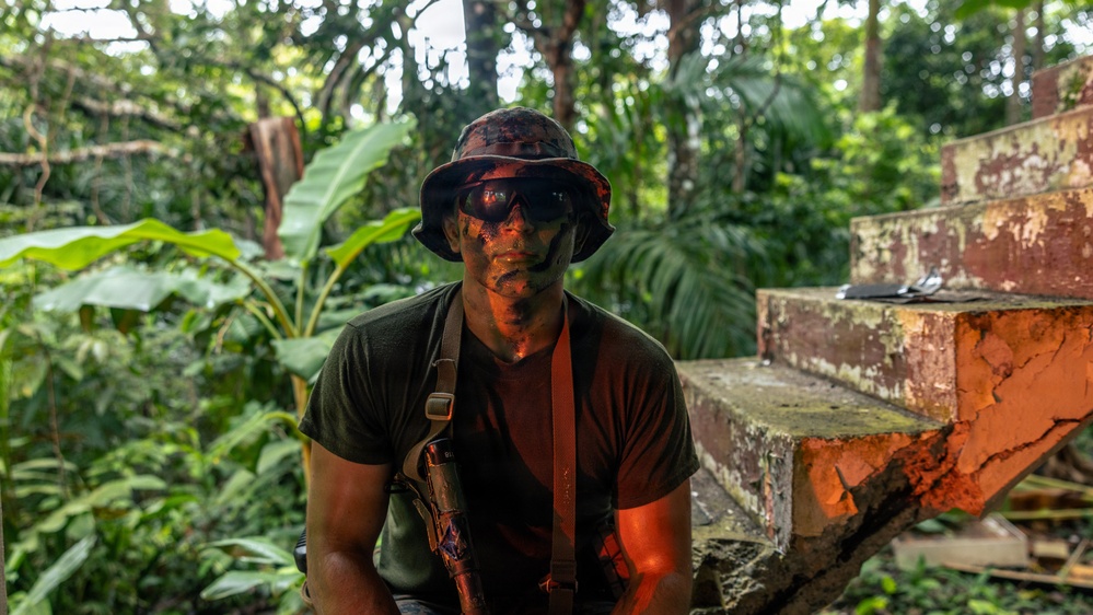 A person wearing camouflage face paint, a camouflage bucket hat, and tactical sunglasses is positioned in a dense tropical jungle environment. They are wearing a dark olive t-shirt with what appears to be a weapon holster or sling across their chest. The setting includes deteriorating brick or concrete structures overgrown with vegetation, suggesting an abandoned or ruined location.