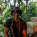 A person wearing camouflage face paint, a camouflage bucket hat, and tactical sunglasses is positioned in a dense tropical jungle environment. They are wearing a dark olive t-shirt with what appears to be a weapon holster or sling across their chest. The setting includes deteriorating brick or concrete structures overgrown with vegetation, suggesting an abandoned or ruined location.