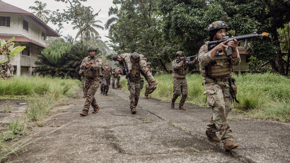 A group of military personnel in multicam/camouflage uniforms are conducting what appears to be a training exercise in a tropical environment. One soldier is performing a fireman's carry on another personnel, simulating a casualty evacuation, while others provide security with raised weapons. The setting includes overgrown vegetation, palm trees, and deteriorating buildings consistent with a Southeast Asian or Pacific Island location.
