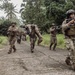 A group of military personnel in multicam/camouflage uniforms are conducting what appears to be a training exercise in a tropical environment. One soldier is performing a fireman's carry on another personnel, simulating a casualty evacuation, while others provide security with raised weapons. The setting includes overgrown vegetation, palm trees, and deteriorating buildings consistent with a Southeast Asian or Pacific Island location.