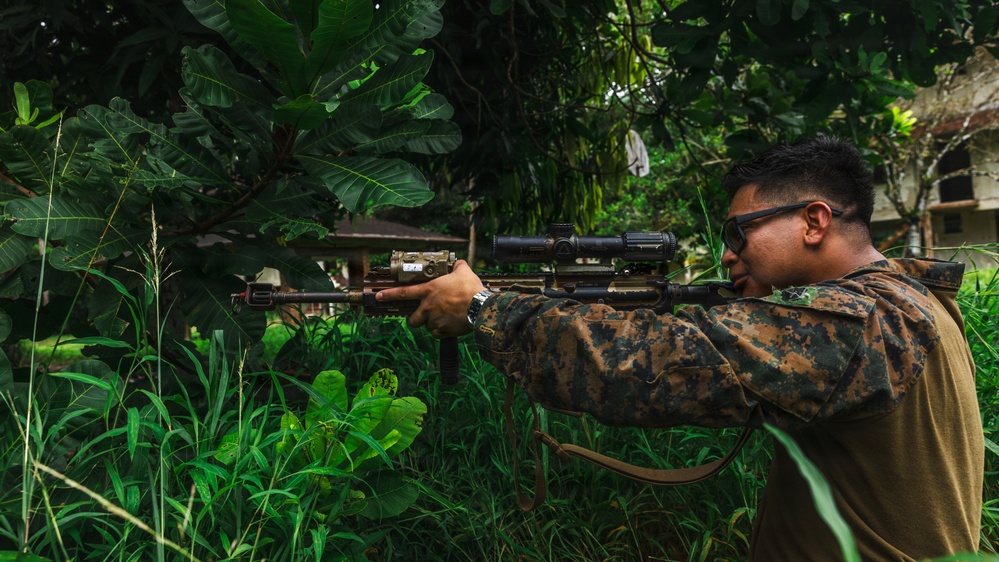 A military service member in camouflage uniform is aiming a rifle equipped with a scope and tactical accessories in a dense, tropical vegetation environment. The individual is positioned in a shooting stance, looking through the optic sight. The setting appears to be a jungle or overgrown area with large-leafed tropical plants and a partially visible structure in the background.