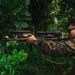 A military service member in camouflage uniform is aiming a rifle equipped with a scope and tactical accessories in a dense, tropical vegetation environment. The individual is positioned in a shooting stance, looking through the optic sight. The setting appears to be a jungle or overgrown area with large-leafed tropical plants and a partially visible structure in the background.