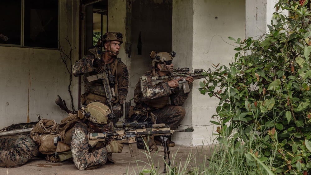 Three military personnel in MARPAT (Marine pattern) camouflage uniforms are positioned at the corner of a deteriorated white concrete building. They appear to be conducting a tactical urban operations exercise or training scenario, with two soldiers in kneeling/crouching positions and one in a prone position. The soldiers are armed and appear to be covering different sectors near a building entrance.