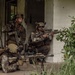 Three military personnel in MARPAT (Marine pattern) camouflage uniforms are positioned at the corner of a deteriorated white concrete building. They appear to be conducting a tactical urban operations exercise or training scenario, with two soldiers in kneeling/crouching positions and one in a prone position. The soldiers are armed and appear to be covering different sectors near a building entrance.