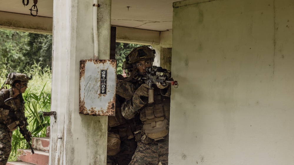 Two military personnel in full combat gear are conducting what appears to be a building clearance or urban warfare training exercise. The primary soldier is positioned at a doorway threshold, aiming a rifle while wearing a helmet with night-vision mount and tactical vest. The setting is a weathered concrete structure surrounded by dense tropical vegetation.