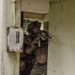 Two military personnel in full combat gear are conducting what appears to be a building clearance or urban warfare training exercise. The primary soldier is positioned at a doorway threshold, aiming a rifle while wearing a helmet with night-vision mount and tactical vest. The setting is a weathered concrete structure surrounded by dense tropical vegetation.