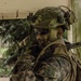 A soldier in full tactical gear is positioned inside or near a partially open structure with tropical vegetation visible in the background. The individual is wearing camouflage uniform, a tactical helmet with mounted accessories, and has green face paint applied, suggesting a field exercise or operational environment. The setting appears to be a tropical or jungle region based on the palm trees and lush greenery visible.