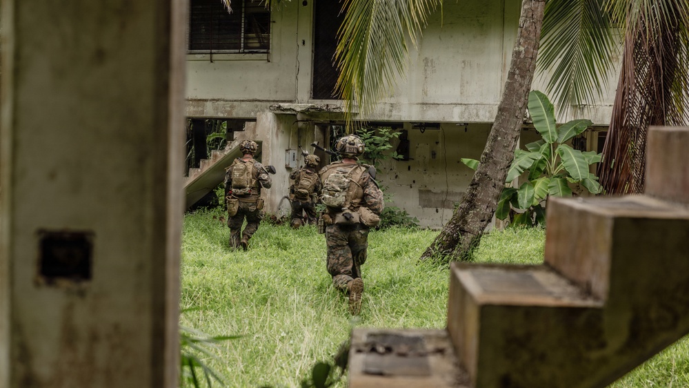The image shows two armed military personnel in full combat gear moving across a grassy area toward a deteriorated concrete building. They appear to be conducting a tactical approach or clearing operation in what looks like a tropical environment, evidenced by palm trees and broad-leafed vegetation. The photo is taken from a partially obscured vantage point, suggesting the photographer is positioned behind cover or a structure.