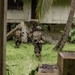 The image shows two armed military personnel in full combat gear moving across a grassy area toward a deteriorated concrete building. They appear to be conducting a tactical approach or clearing operation in what looks like a tropical environment, evidenced by palm trees and broad-leafed vegetation. The photo is taken from a partially obscured vantage point, suggesting the photographer is positioned behind cover or a structure.
