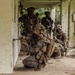 A group of military personnel in full combat gear are conducting what appears to be a tactical building clearance or urban warfare training exercise. The soldiers are positioned in a stacked formation along a corridor of a deteriorated structure, with the lead soldier kneeling and aiming a rifle around a corner. The setting appears to be an abandoned or dilapidated building surrounded by dense tropical vegetation.