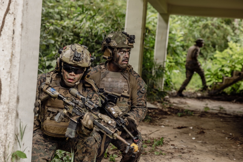 Two heavily equipped military personnel in MARPAT (Marine pattern) camouflage uniforms are positioned against a concrete structure in a jungle or tropical environment, appearing to conduct a tactical operation or training exercise. A third individual in camouflage is visible in the background walking along a path. The foreground soldier is armed with what appears to be an M4/M16-variant rifle with multiple attachments.