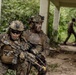 Two heavily equipped military personnel in MARPAT (Marine pattern) camouflage uniforms are positioned against a concrete structure in a jungle or tropical environment, appearing to conduct a tactical operation or training exercise. A third individual in camouflage is visible in the background walking along a path. The foreground soldier is armed with what appears to be an M4/M16-variant rifle with multiple attachments.