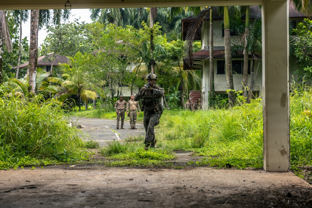 The image shows military or law enforcement personnel conducting what appears to be a patrol or training exercise in a tropical, overgrown compound with abandoned or semi-abandoned buildings. One heavily equipped soldier in full combat gear and helmet moves toward the camera while two others in lighter tactical clothing stand further back on a paved path. The lush tropical vegetation, palm trees, and deteriorating structures suggest a Southeast Asian or Pacific Island location.