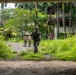 The image shows military or law enforcement personnel conducting what appears to be a patrol or training exercise in a tropical, overgrown compound with abandoned or semi-abandoned buildings. One heavily equipped soldier in full combat gear and helmet moves toward the camera while two others in lighter tactical clothing stand further back on a paved path. The lush tropical vegetation, palm trees, and deteriorating structures suggest a Southeast Asian or Pacific Island location.