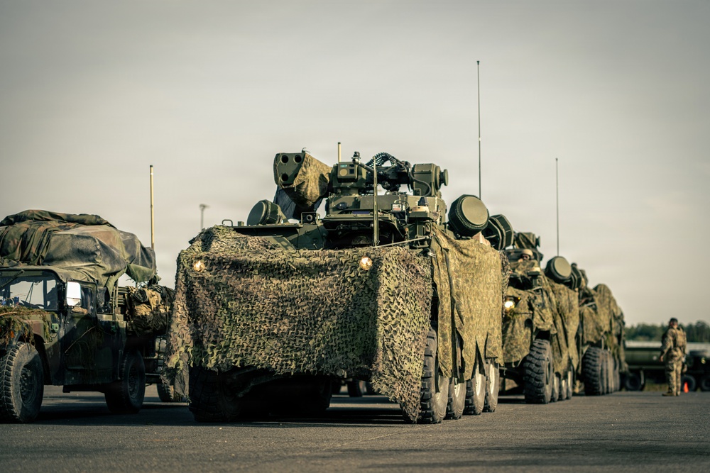 A military convoy of wheeled armored vehicles is staged on a paved surface, likely a road or airfield tarmac. The vehicles are draped in camouflage netting and appear to be equipped with weapon systems and communication antennas. At least one soldier is visible in the background on the right side of the frame.