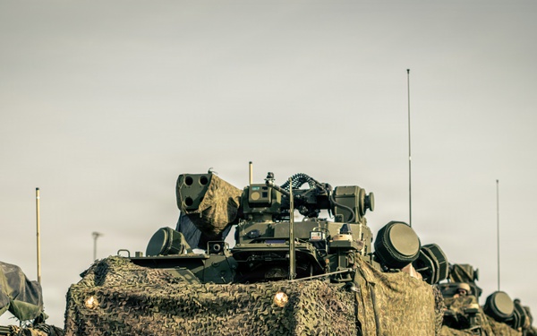 A military convoy of wheeled armored vehicles is staged on a paved surface, likely a road or airfield tarmac. The vehicles are draped in camouflage netting and appear to be equipped with weapon systems and communication antennas. At least one soldier is visible in the background on the right side of the frame.