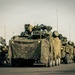 A military convoy of wheeled armored vehicles is staged on a paved surface, likely a road or airfield tarmac. The vehicles are draped in camouflage netting and appear to be equipped with weapon systems and communication antennas. At least one soldier is visible in the background on the right side of the frame.