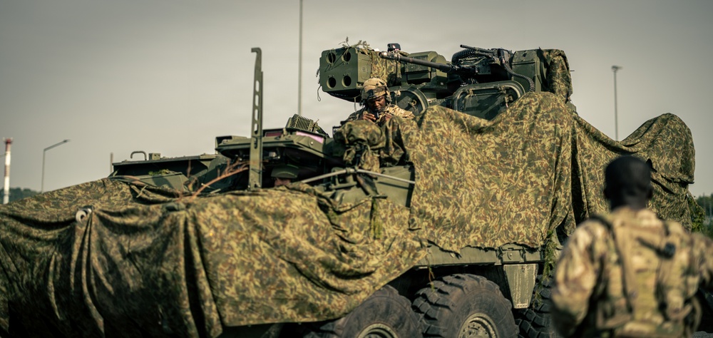 The image shows military personnel operating what appears to be a Stryker armored fighting vehicle draped with camouflage netting. At least two soldiers in combat gear and helmets are visible, one positioned in the vehicle's turret hatch. The scene appears to be a military exercise or operational deployment in an open area.