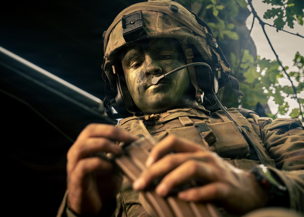 A soldier in full tactical gear is photographed from a low angle, with face camouflage paint applied in dark green and black tones. The soldier is handling what appears to be a firearm magazine or weapon component with both hands. The image is dramatically lit with trees visible in the background, suggesting an outdoor field exercise or operational environment.