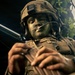 A soldier in full tactical gear is photographed from a low angle, with face camouflage paint applied in dark green and black tones. The soldier is handling what appears to be a firearm magazine or weapon component with both hands. The image is dramatically lit with trees visible in the background, suggesting an outdoor field exercise or operational environment.