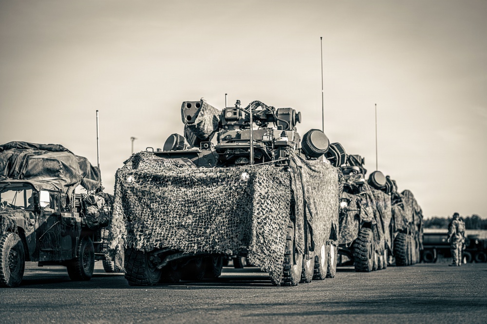 A military convoy of wheeled armored vehicles is staged on a paved surface, likely an airfield or staging area. The lead vehicle is draped in camouflage netting and equipped with a remote weapon station or turret on top. At least one soldier is visible in the background, and a HMMWV (Humvee) is present on the left side of the formation.
