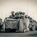 A military convoy of wheeled armored vehicles is staged on a paved surface, likely an airfield or staging area. The lead vehicle is draped in camouflage netting and equipped with a remote weapon station or turret on top. At least one soldier is visible in the background, and a HMMWV (Humvee) is present on the left side of the formation.
