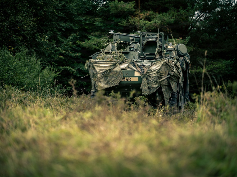 A military armored vehicle, partially concealed with camouflage netting and natural vegetation, is positioned at the edge of a treeline in a field setting. The vehicle bears visible text reading 'WAR' on its hull and is accompanied by at least one soldier in camouflage gear standing beside it. The scene appears to depict a tactical concealment or observation position during a military exercise or operation.