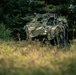 A military armored vehicle, partially concealed with camouflage netting and natural vegetation, is positioned at the edge of a treeline in a field setting. The vehicle bears visible text reading 'WAR' on its hull and is accompanied by at least one soldier in camouflage gear standing beside it. The scene appears to depict a tactical concealment or observation position during a military exercise or operation.
