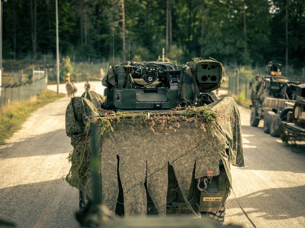 A military convoy is moving along a gravel road through a forested area. The lead vehicle is heavily draped with camouflage netting and natural vegetation for concealment, with military equipment visible on top. At least two additional military vehicles are visible in the convoy, along with personnel on foot in the background.