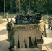 A military convoy is moving along a gravel road through a forested area. The lead vehicle is heavily draped with camouflage netting and natural vegetation for concealment, with military equipment visible on top. At least two additional military vehicles are visible in the convoy, along with personnel on foot in the background.