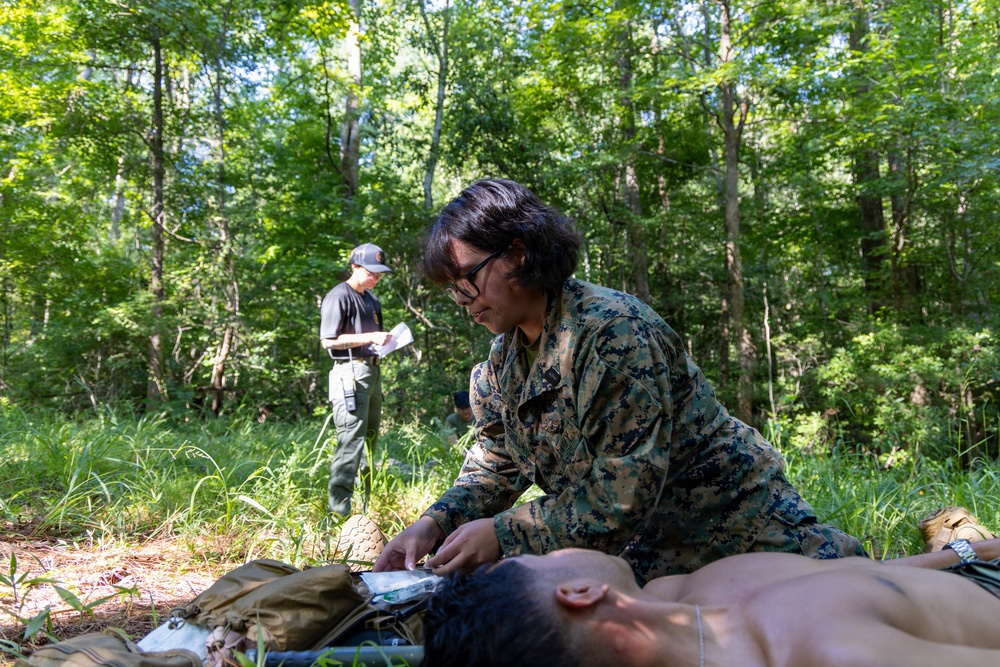 A military service member in MARPAT digital camouflage uniform is performing field medical treatment on a casualty lying on the ground in a wooded outdoor setting. A second individual in civilian-style clothing and a cap stands in the background appearing to observe and take notes, suggesting this is a training evaluation scenario. The setting is a sunlit forest clearing with dense green vegetation.