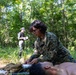 A military service member in MARPAT digital camouflage uniform is performing field medical treatment on a casualty lying on the ground in a wooded outdoor setting. A second individual in civilian-style clothing and a cap stands in the background appearing to observe and take notes, suggesting this is a training evaluation scenario. The setting is a sunlit forest clearing with dense green vegetation.