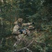 Two individuals in full camouflage military-style gear are positioned in a dense forest setting. The foreground figure is crouching and pointing in a direction while holding an assault-style rifle with an optic sight. A second figure in a boonie hat is visible in the background, also holding a firearm in a prone or low position.