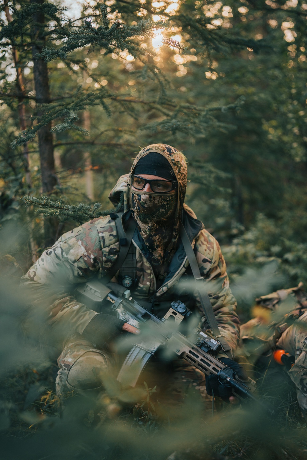 A person in full camouflage tactical gear is crouched low in a forested environment, holding a rifle with accessories. They are wearing a camouflage balaclava, protective eyewear, and a hooded jacket, blending into the woodland surroundings. The setting and orange-tipped barrel visible on a second weapon suggest this is an airsoft or milsim (military simulation) activity.