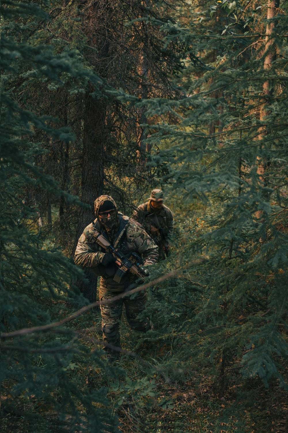 Two individuals in full camouflage tactical gear are moving through a dense coniferous forest. The lead figure is carrying what appears to be an assault-style rifle and is wearing a balaclava, hood, and protective eyewear. The image appears to depict a military patrol, tactical training exercise, or airsoft/milsim activity.