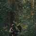 Two individuals in full camouflage tactical gear are moving through a dense coniferous forest. The lead figure is carrying what appears to be an assault-style rifle and is wearing a balaclava, hood, and protective eyewear. The image appears to depict a military patrol, tactical training exercise, or airsoft/milsim activity.