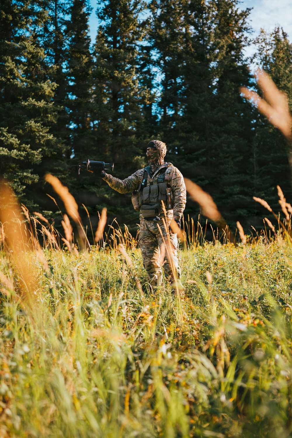 A person dressed in full camouflage gear stands in a sunlit meadow surrounded by tall conifer trees. They are holding a small device in one outstretched hand, wearing a face covering, cap, and tactical vest with pouches. The scene appears to be an outdoor field setting, likely during early morning or late afternoon based on the warm directional light.