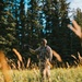 A person dressed in full camouflage gear stands in a sunlit meadow surrounded by tall conifer trees. They are holding a small device in one outstretched hand, wearing a face covering, cap, and tactical vest with pouches. The scene appears to be an outdoor field setting, likely during early morning or late afternoon based on the warm directional light.