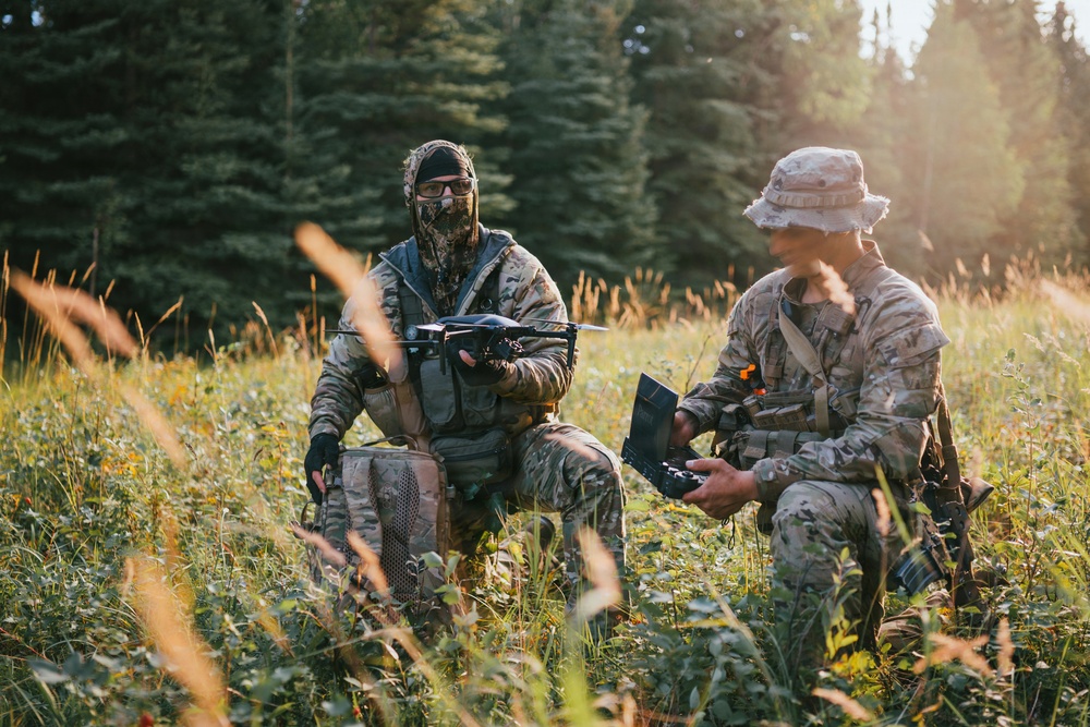 Two individuals in military-style camouflage uniforms are positioned in an open field surrounded by coniferous trees. One person is holding a small quadcopter drone while the other holds what appears to be a remote control unit or tablet. The scene appears to depict drone operation or preparation in an outdoor tactical setting.