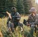 Two individuals in military-style camouflage uniforms are positioned in an open field surrounded by coniferous trees. One person is holding a small quadcopter drone while the other holds what appears to be a remote control unit or tablet. The scene appears to depict drone operation or preparation in an outdoor tactical setting.