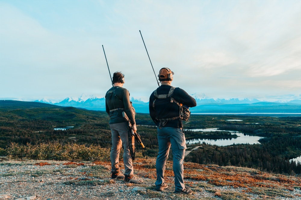 Two individuals stand on an elevated rocky ridge overlooking a vast wilderness landscape featuring forests, lakes, and snow-capped mountains in the distance. Both are equipped with backpacks fitted with long radio/telemetry antennas and appear to be engaged in wildlife tracking or surveying activities. The scene suggests a remote backcountry environment, likely in Alaska or a similar subarctic region.