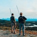 Two individuals stand on an elevated rocky ridge overlooking a vast wilderness landscape featuring forests, lakes, and snow-capped mountains in the distance. Both are equipped with backpacks fitted with long radio/telemetry antennas and appear to be engaged in wildlife tracking or surveying activities. The scene suggests a remote backcountry environment, likely in Alaska or a similar subarctic region.
