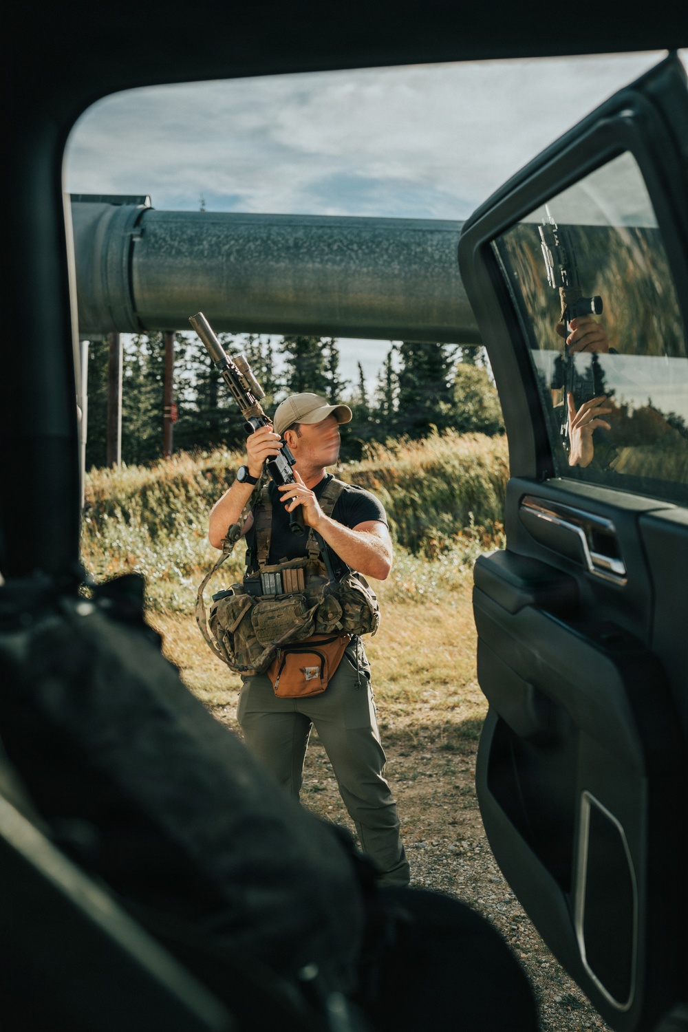 A person stands outside a large truck or SUV in an outdoor rural setting, holding a suppressed rifle in a ready position while wearing tactical gear. The image is framed through the open door of the vehicle, with a large pipeline visible in the background along with conifer trees and open grassland. The scene appears consistent with a hunting or tactical training context in a remote area.