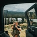 A person stands outside a large truck or SUV in an outdoor rural setting, holding a suppressed rifle in a ready position while wearing tactical gear. The image is framed through the open door of the vehicle, with a large pipeline visible in the background along with conifer trees and open grassland. The scene appears consistent with a hunting or tactical training context in a remote area.