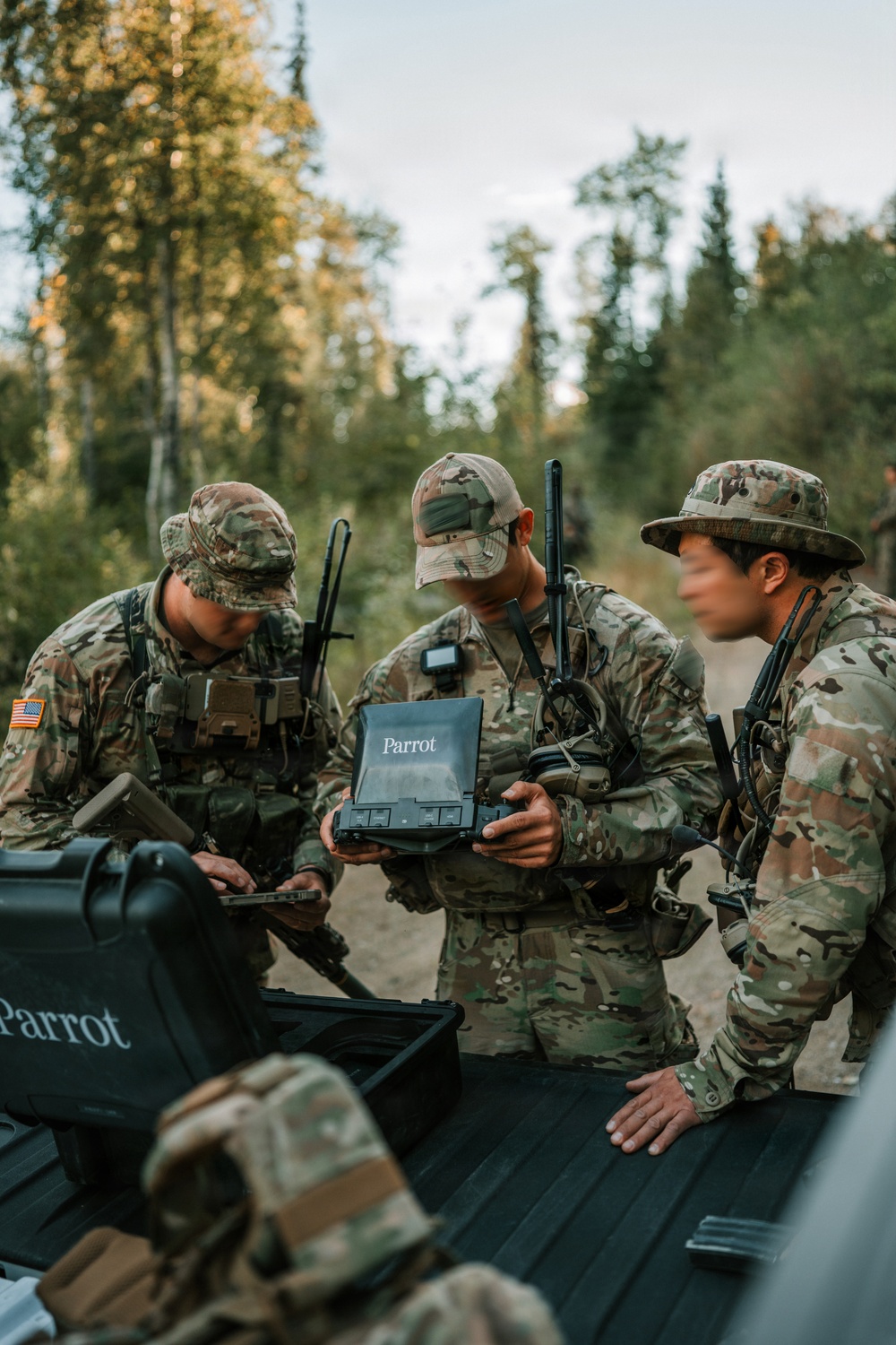 Three U.S. Army soldiers in MultiCam/OCP camouflage uniforms are gathered around the bed of a pickup truck in a forested outdoor environment. They are operating Parrot-branded drone equipment, including a rugged tablet controller and a carrying case. The setting appears to be a field training exercise or operational deployment.