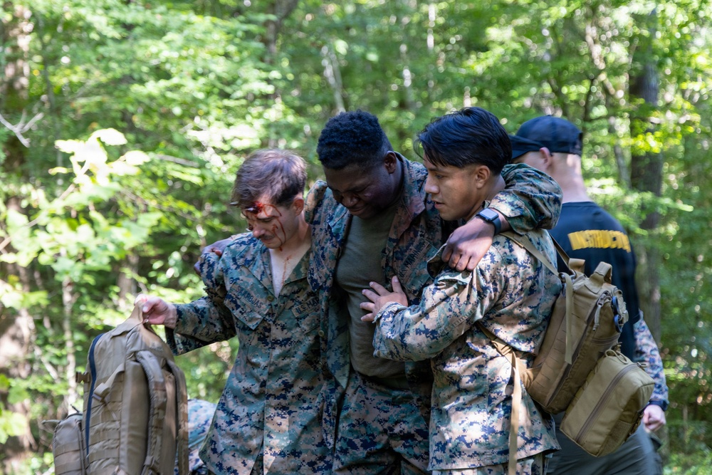 The image shows what appears to be a military training exercise in a wooded outdoor setting. Two personnel in U.S. Marine Corps MARPAT camouflage uniforms are assisting a third individual who appears to have simulated facial injuries (moulage makeup). A fourth individual in a dark uniform with a cap is visible in the background.
