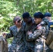 The image shows what appears to be a military training exercise in a wooded outdoor setting. Two personnel in U.S. Marine Corps MARPAT camouflage uniforms are assisting a third individual who appears to have simulated facial injuries (moulage makeup). A fourth individual in a dark uniform with a cap is visible in the background.